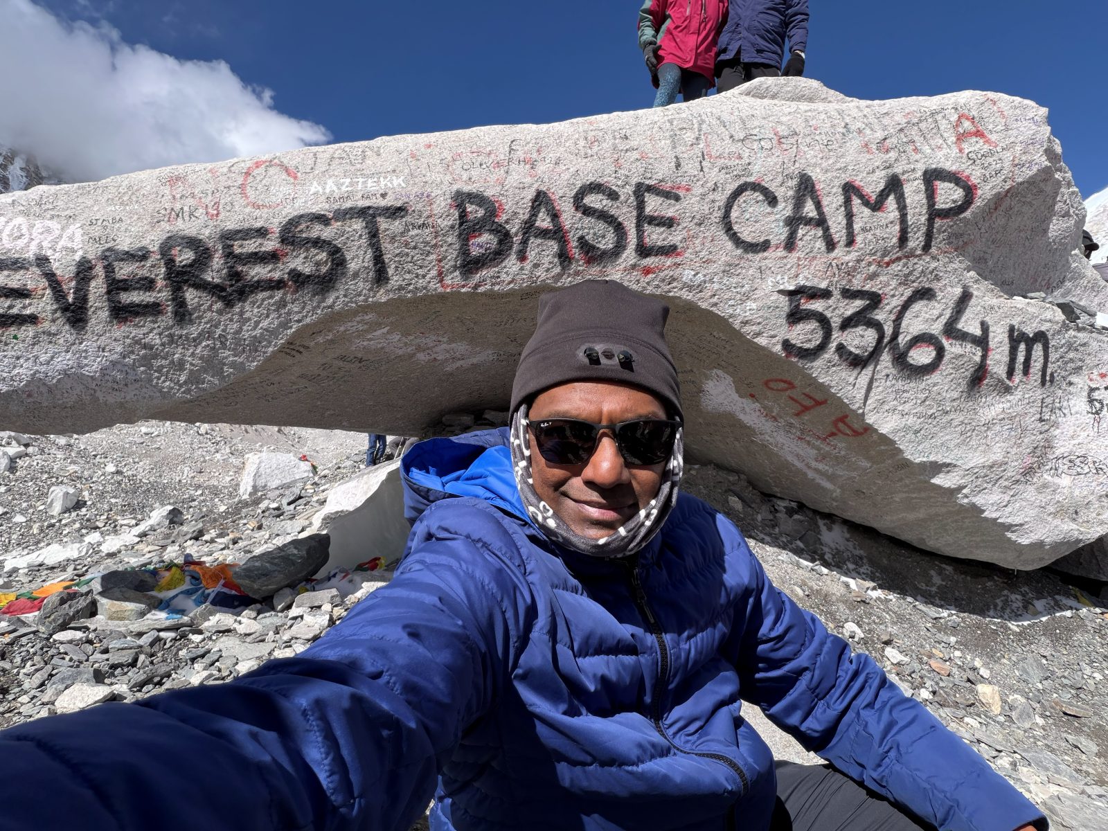 Anand in a blue down jacket and sunglasses in front of the painted Everest Base Camp boulder marked 5364 m